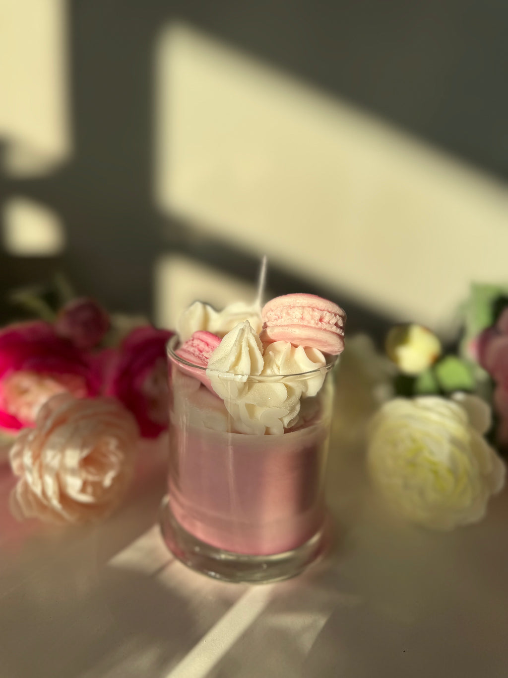 Pink dessert with whipped cream and macarons in a glass, surrounded by flowers.