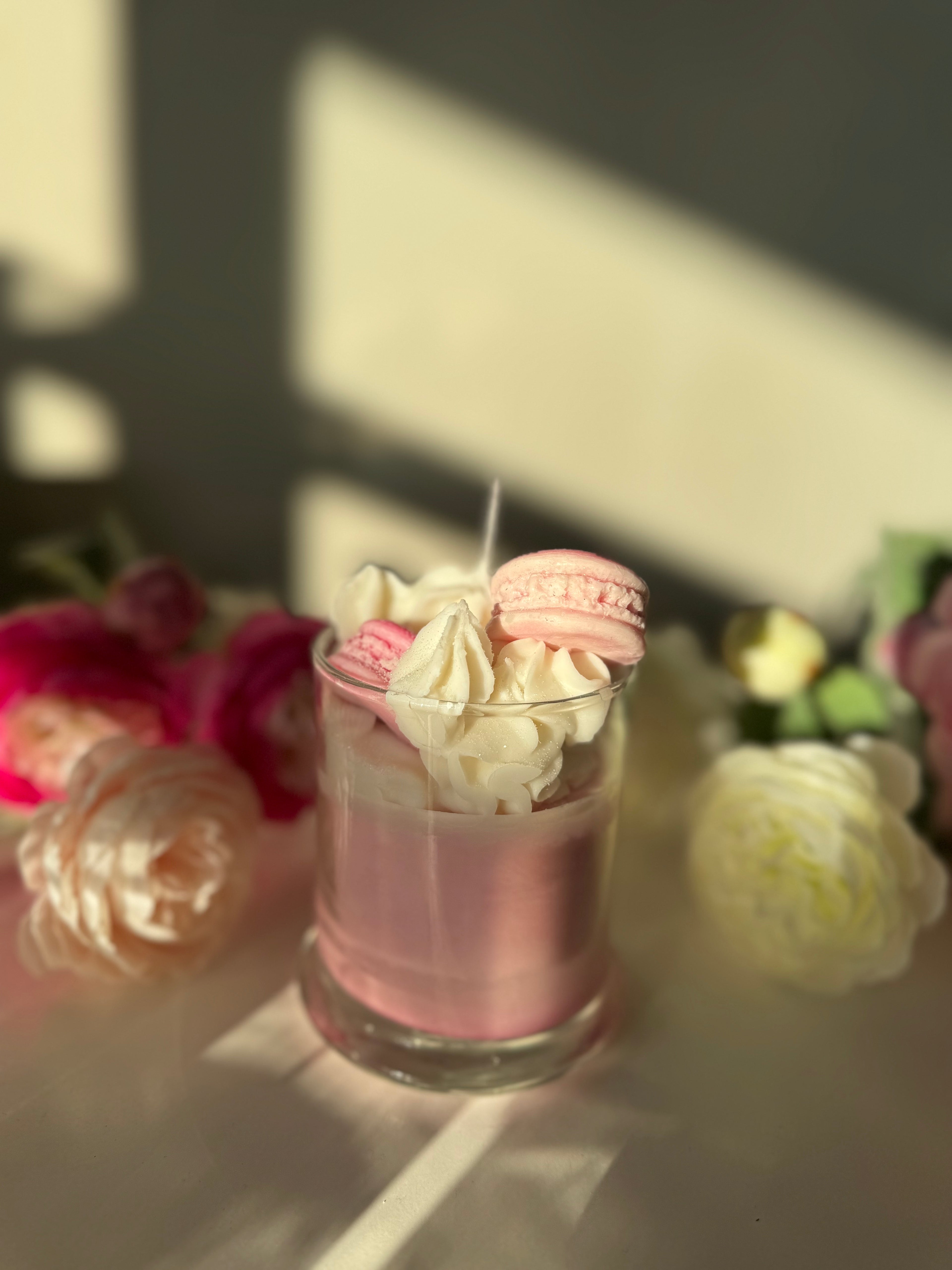 Pink dessert with whipped cream and macarons in a glass, surrounded by flowers.