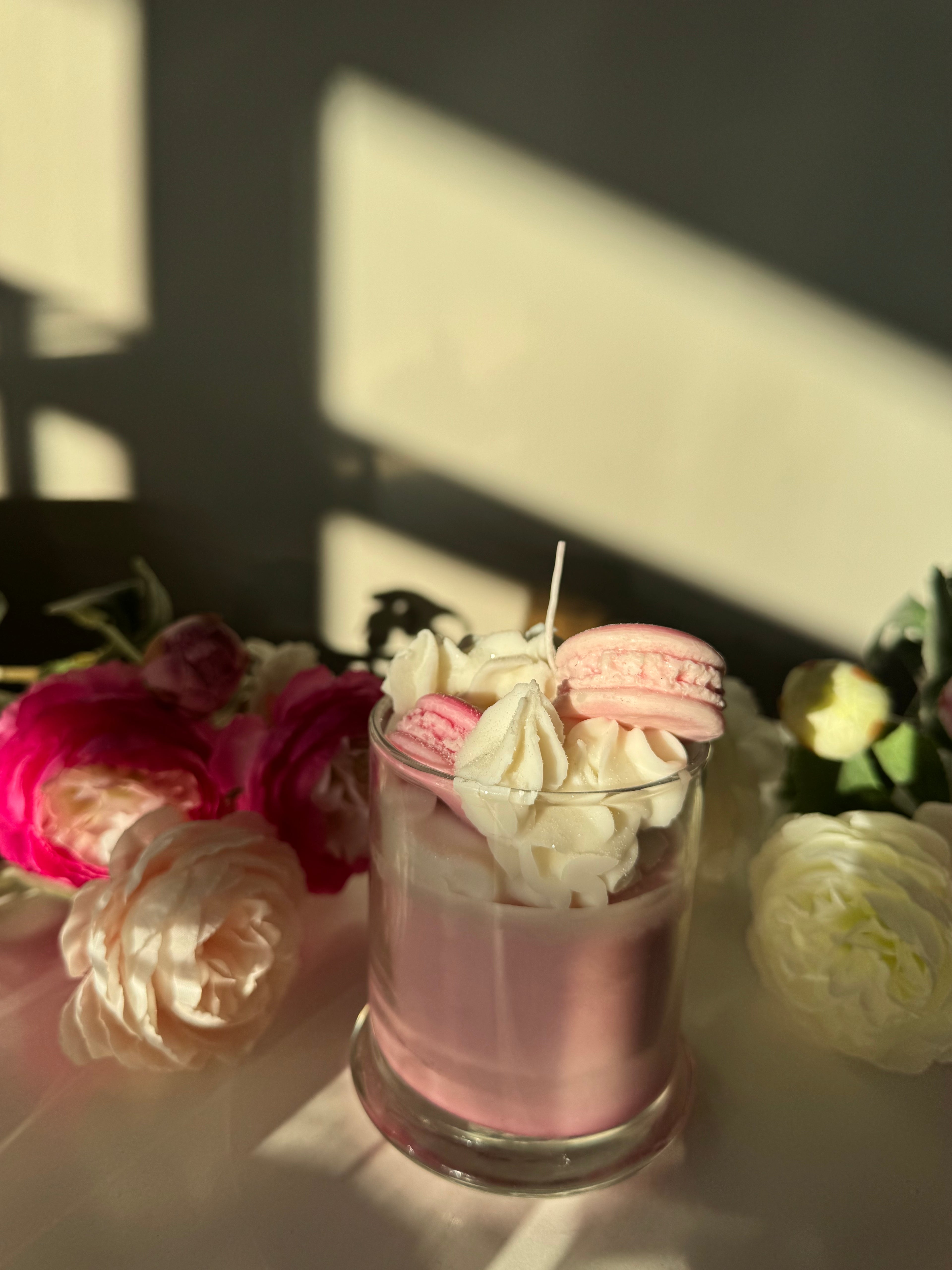 Pink macaron, white cream, and pink candle on a reflective surface with flowers in the background